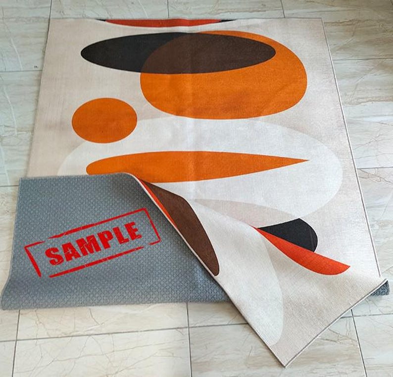May include: A rectangular area rug featuring an abstract pattern of overlapping orange, brown, and white circular and oval shapes. The rug is partially folded to reveal a gray, textured, non-slip backing with the red text SAMPLE printed on it.