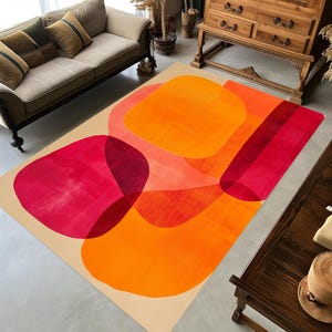 Op de afbeelding: Een rechthoekig vloerkleed met een beige achtergrond en grote, overlappende abstracte vormen in levendige tinten oranje, felrood en diep magenta. Het ontwerp bevat afgeronde vormen en rechte banen die een gelaagd effect creëren.