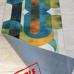 May include: A rectangular area rug with an abstract geometric design in shades of green, blue, and orange on a white background. The rug's underside is visible, showing a blue patterned backing. A red "SAMPLE" stamp is visible.