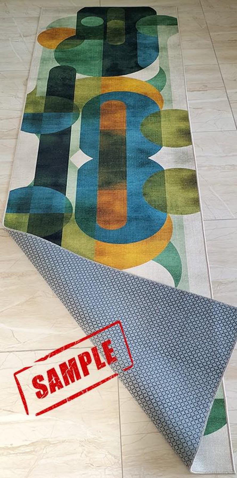 May include: A rectangular area rug with an abstract geometric design in shades of green, blue, and orange on a white background. The rug's underside is visible, showing a blue patterned backing. A red "SAMPLE" stamp is visible.