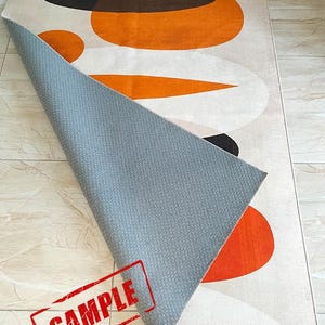 May include: A rectangular area rug featuring a pattern of large orange, brown, and white oval shapes. The rug is folded over to reveal a gray, textured, non-slip backing. The word SAMPLE is printed in red ink across the bottom left corner.
