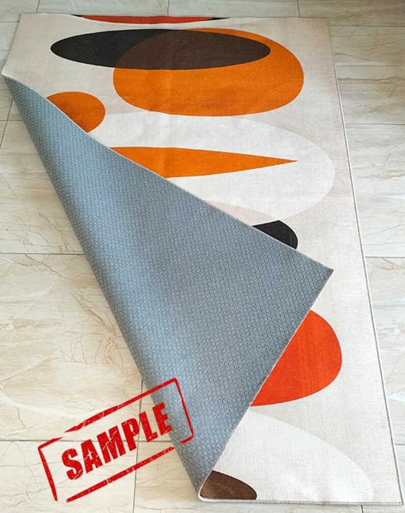 May include: A rectangular area rug featuring a pattern of large orange, brown, and white oval shapes. The rug is folded over to reveal a gray, textured, non-slip backing. The word SAMPLE is printed in red ink across the bottom left corner.