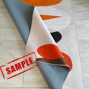 May include: A rectangular area rug featuring a geometric pattern of large orange, brown, and off-white circles and shapes. The rug is folded over to reveal a gray, textured, non-slip backing. The word SAMPLE is printed in red on the lower left corner.