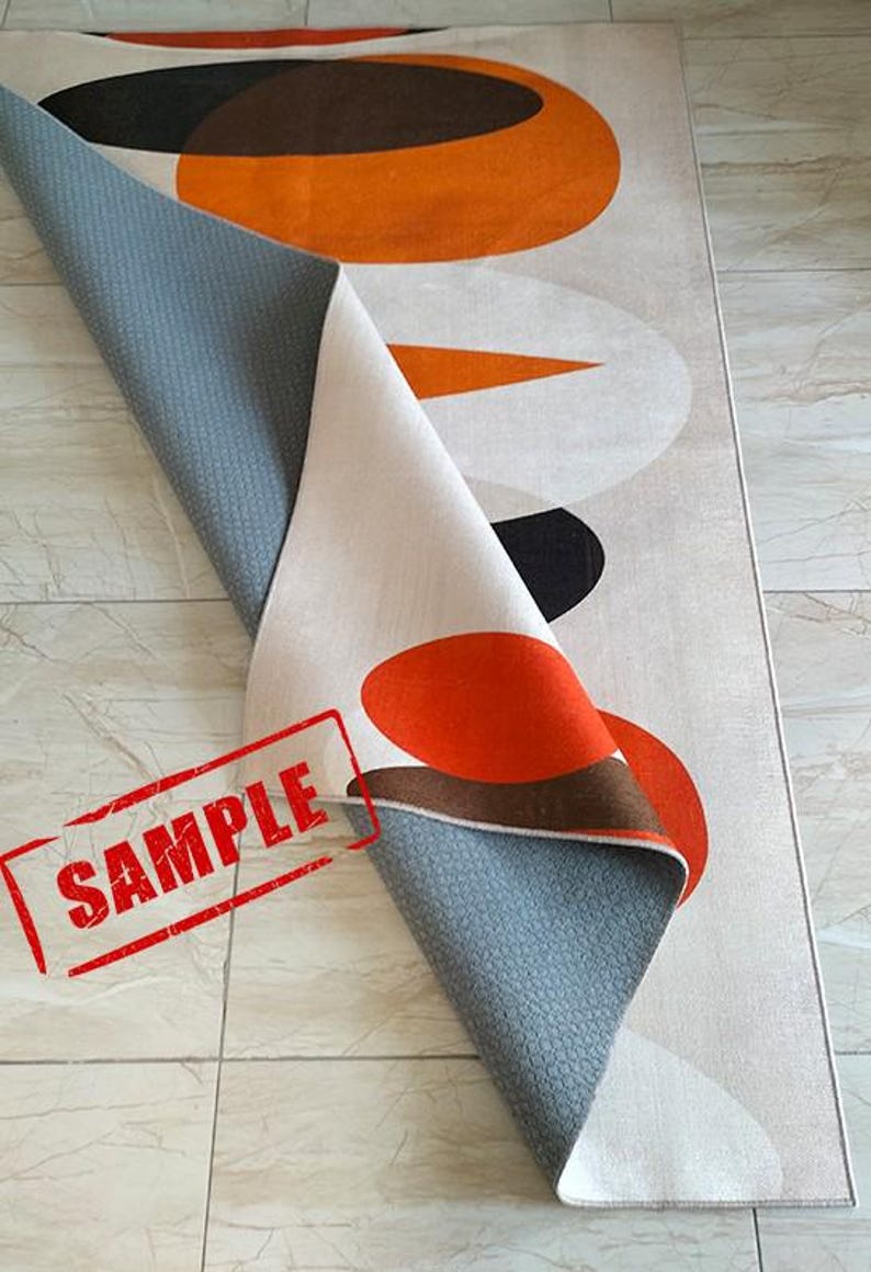 May include: A rectangular area rug featuring a geometric pattern of large orange, brown, and off-white circles and shapes. The rug is folded over to reveal a gray, textured, non-slip backing. The word SAMPLE is printed in red on the lower left corner.