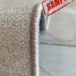 May include: A beige rug with a thick, tightly woven overlocked edge finish. The rug features a textured, short-pile surface and is folded to display the side profile. The word SAMPLE is printed in red ink across the upper right corner.