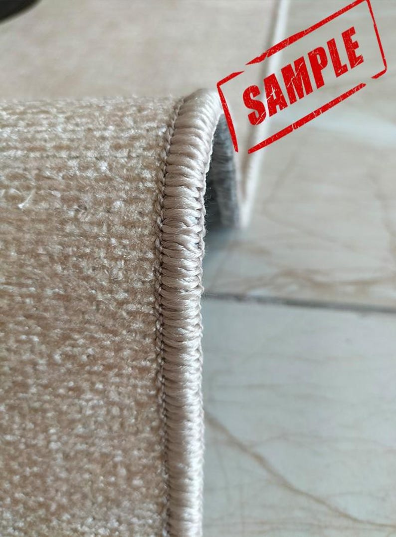 May include: A beige rug with a thick, tightly woven overlocked edge finish. The rug features a textured, short-pile surface and is folded to display the side profile. The word SAMPLE is printed in red ink across the upper right corner.