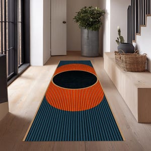 May include: A rectangular rug with a geometric design. The rug features a large black oval in the center, surrounded by a vibrant orange semi-circle and a blue striped background. The rug has a beige border and is placed on a light wood floor.