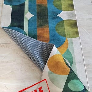 May include: A rectangular rug with an abstract design featuring overlapping shapes in shades of green, blue, and gold. The rug is partially rolled up, revealing a gray patterned underside. The word "SAMPLE" is stamped in red.