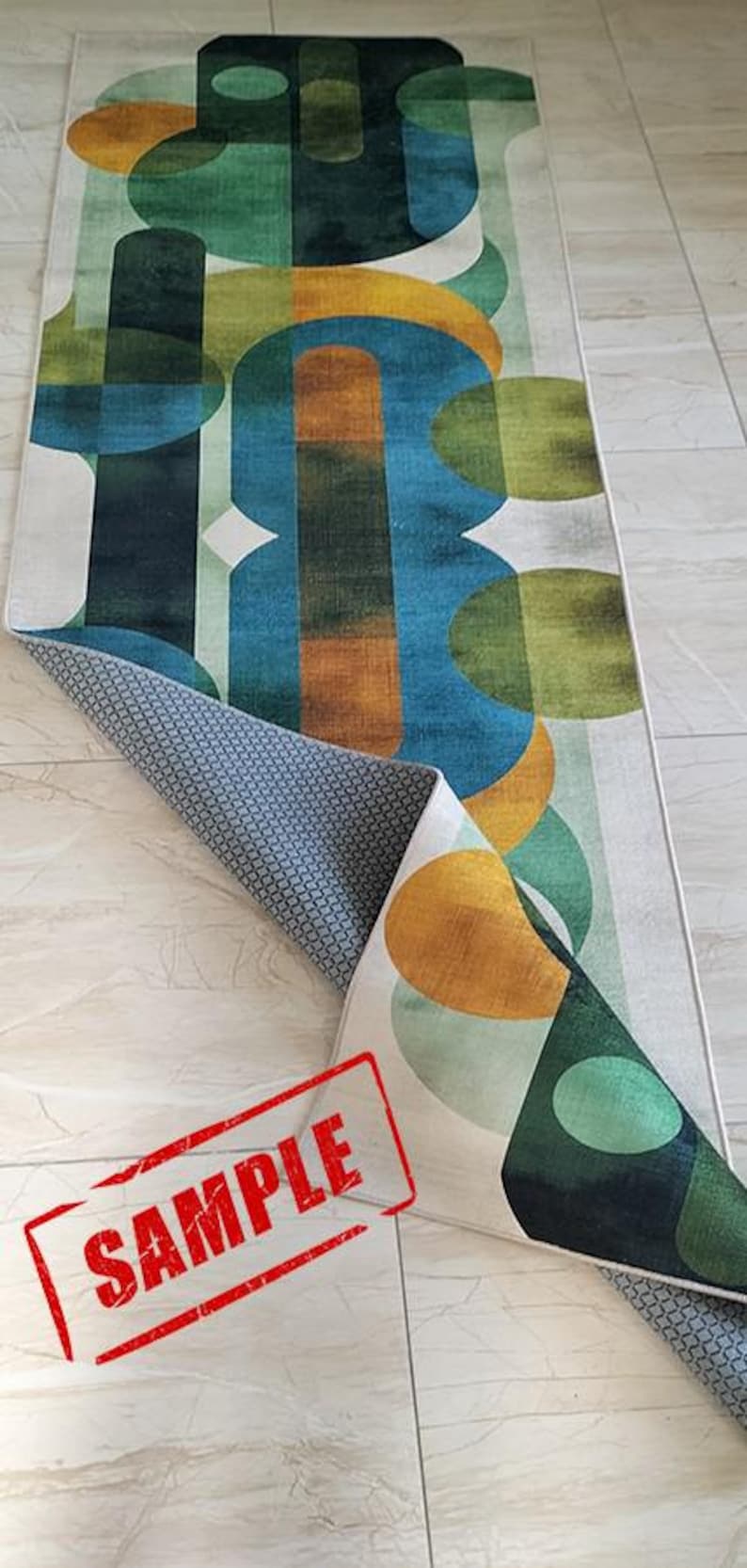 May include: A rectangular rug with an abstract design featuring overlapping shapes in shades of green, blue, and gold. The rug is partially rolled up, revealing a gray patterned underside. The word "SAMPLE" is stamped in red.