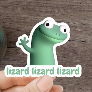 May include: A green lizard sticker with the text "lizard lizard lizard" is held by a hand. The sticker features a cartoon lizard waving. A glass jar and a spiral-bound notebook are also visible on a wooden surface.