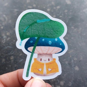 Op de afbeelding: Een sticker van een cartoon paddenstoel die een gele regenjas draagt en een groene bladerenparaplu vasthoudt. De paddenstoel heeft een blauwe dop met witte stippen en een roze steel.