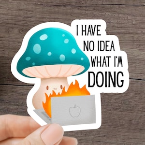 Puede incluir: Una pegatina con un hongo de dibujos animados con una gorra turquesa y puntos blancos, sosteniendo un portátil en llamas. El texto "I HAVE NO IDEA WHAT I'M DOING" está en negro. La pegatina está sujeta por una mano.
