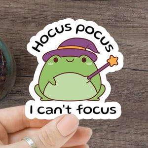Può includere: Una rana verde con un cappello da strega viola e un bastone magico. La rana è circondata da un cerchio bianco con il testo "Hocus Pocus I can't focus".