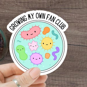 Puede incluir: Una pegatina blanca con el texto "GROWING MY OWN FAN CLUB" en un diseño circular. La pegatina presenta microbios de dibujos animados en varios colores, incluyendo rosa, amarillo, verde y morado, sobre un fondo azul claro. También se ven un tarro de cristal y un cuaderno de espiral.