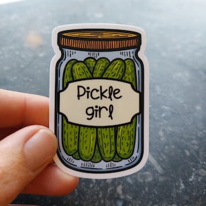 May include: A white sticker featuring a cartoon illustration of a glass jar filled with green pickles. The jar has a brown lid and a white label that reads "Pickle girl".
