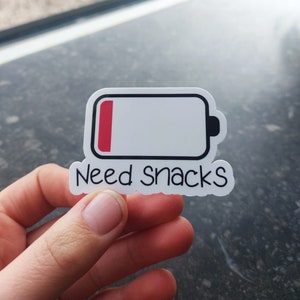 Op de afbeelding: Witte sticker met een rood en zwart batterijpictogram dat bijna leeg is. De tekst "Need snacks" staat onder het batterijpictogram.