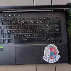 May include: A black laptop computer with a sticker on the right side of the touchpad. The sticker features a brain with flowers and the text "Be kind to your mind".