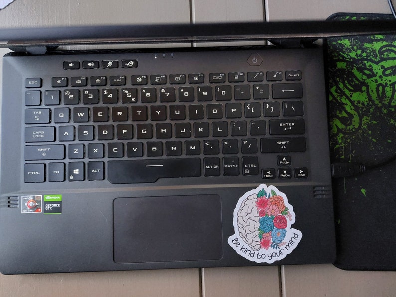 May include: A black laptop computer with a sticker on the right side of the touchpad. The sticker features a brain with flowers and the text "Be kind to your mind".