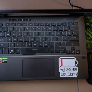 May include: A black laptop with a sticker that says "My social battery" and a low battery icon. The laptop has a black keyboard and a touchpad.