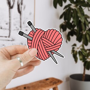 May include: A pink heart-shaped yarn ball with two knitting needles sticking out of it. The yarn ball is held in a hand.