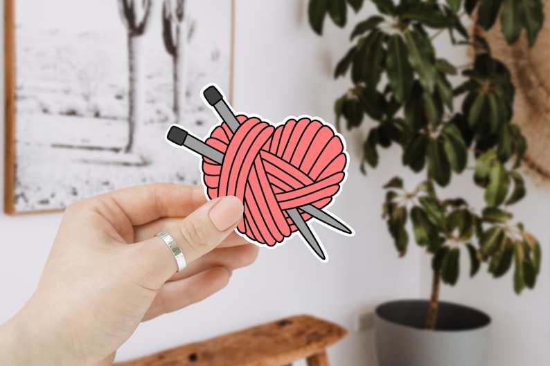 May include: A pink heart-shaped yarn ball with two knitting needles sticking out of it. The yarn ball is held in a hand.