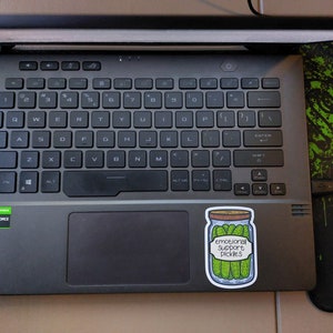 May include: A sticker on a laptop computer with a drawing of a jar of pickles. The jar has a label that reads "Emotional Support Pickles".
