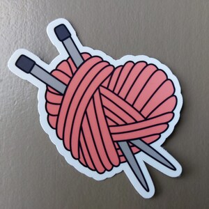 May include: A pink yarn ball with two knitting needles crossed through it, forming a heart shape.