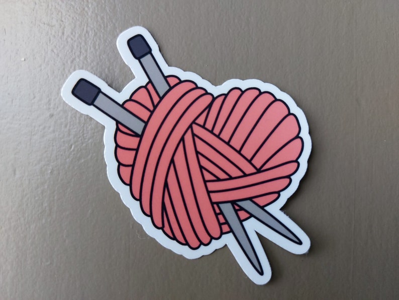May include: A pink yarn ball with two knitting needles crossed through it, forming a heart shape.