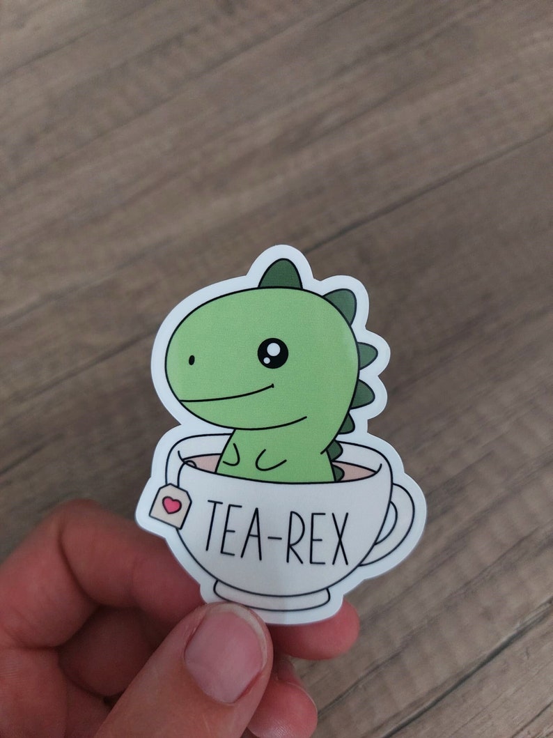 May include: A sticker featuring a green dinosaur in a white teacup. The teacup has the words "TEA-REX" printed on it. The dinosaur has a friendly expression and is surrounded by a white border.
