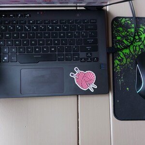 May include: A black laptop computer with a pink yarn ball and knitting needles sticker on the lid. A black wireless mouse with a green light is on a black and green mousepad.