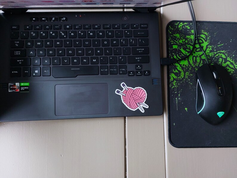 May include: A black laptop computer with a pink yarn ball and knitting needles sticker on the lid. A black wireless mouse with a green light is on a black and green mousepad.