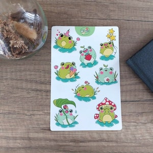 Op de afbeelding: Een vel stickers met schattige cartoon kikkers in verschillende poses. De kikkers zijn groen met zwarte ogen en zijn omringd door bloemen, paddenstoelen en andere natuurthema's. De stickers zijn perfect om een ​​tikje whimsy toe te voegen aan dagboeken, laptops of waterflessen.