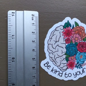 May include: A white sticker with a gray brain outline and colorful flowers. The text "Be kind to your mind" is written in a white cursive font.