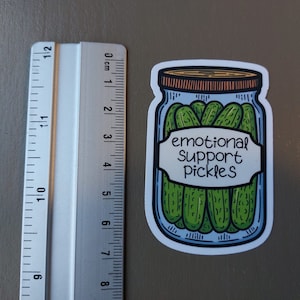 May include: A white sticker with a cartoon illustration of a glass jar filled with green pickles. The jar has a brown lid and a white label that reads "emotional support pickles".