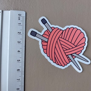 May include: A pink yarn ball with two knitting needles crossed over it. The yarn ball is in the shape of a heart.