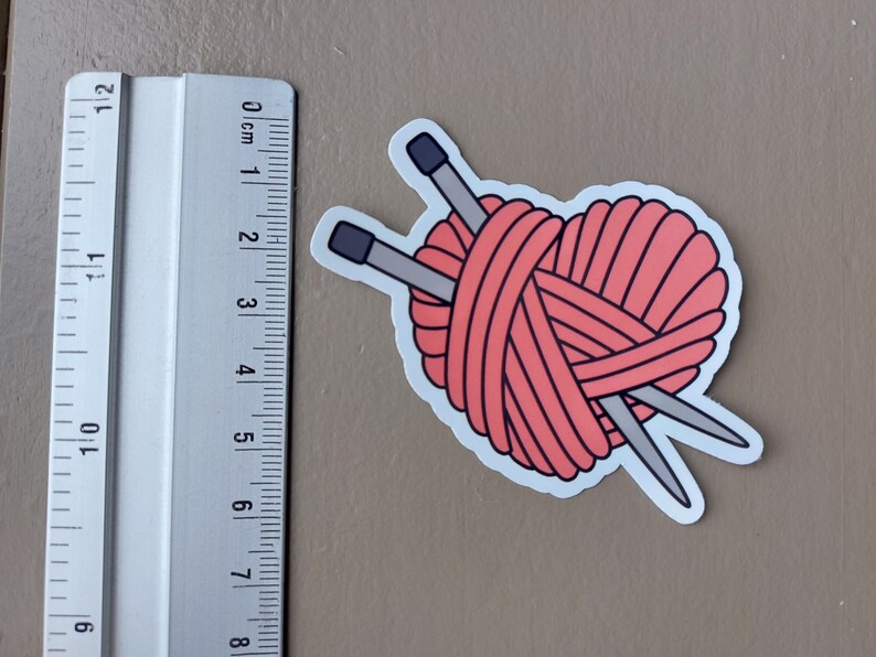 May include: A pink yarn ball with two knitting needles crossed over it. The yarn ball is in the shape of a heart.