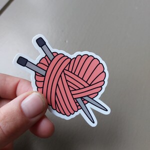 May include: A pink yarn ball in the shape of a heart with two knitting needles crossing through it.