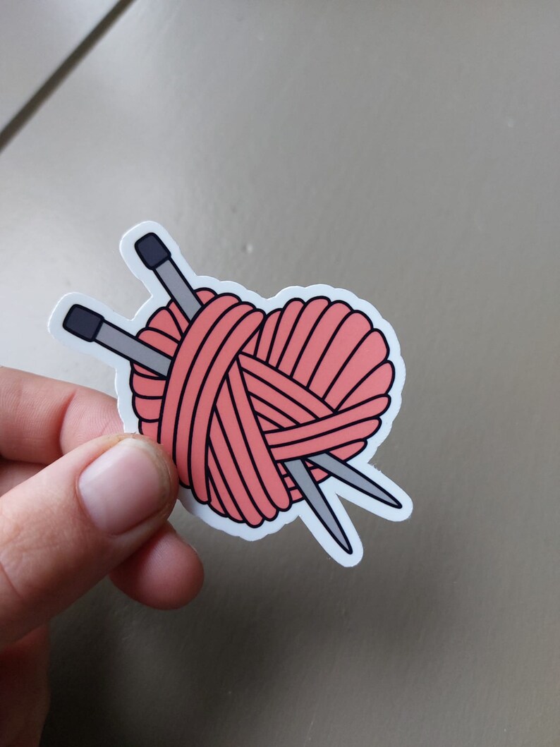 May include: A pink yarn ball in the shape of a heart with two knitting needles crossing through it.