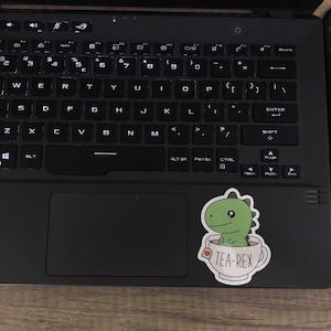 May include: A black laptop with a keyboard and a sticker of a green dinosaur in a teacup that says "TEA-REX". The laptop is on a wooden surface, next to a black and green mousepad.
