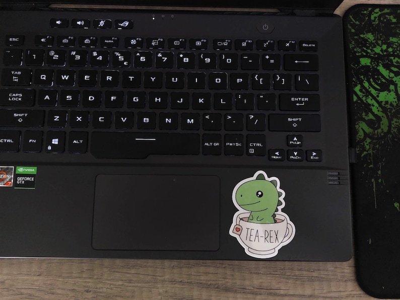 May include: A black laptop with a keyboard and a sticker of a green dinosaur in a teacup that says "TEA-REX". The laptop is on a wooden surface, next to a black and green mousepad.