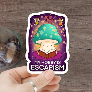 Op de afbeelding: Een grillige sticker met een lachende paddenstoel die een boek leest, met een turquoise kap en een paarse achtergrond. De tekst "MY HOBBY IS ESCAPISM" staat eronder, met feeën en een draak erboven.
