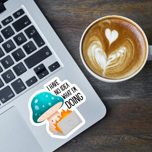 May include: A laptop with a sticker featuring a mushroom and a burning laptop, with the text "I have no idea what I'm doing." A cup of coffee with latte art is next to the laptop.