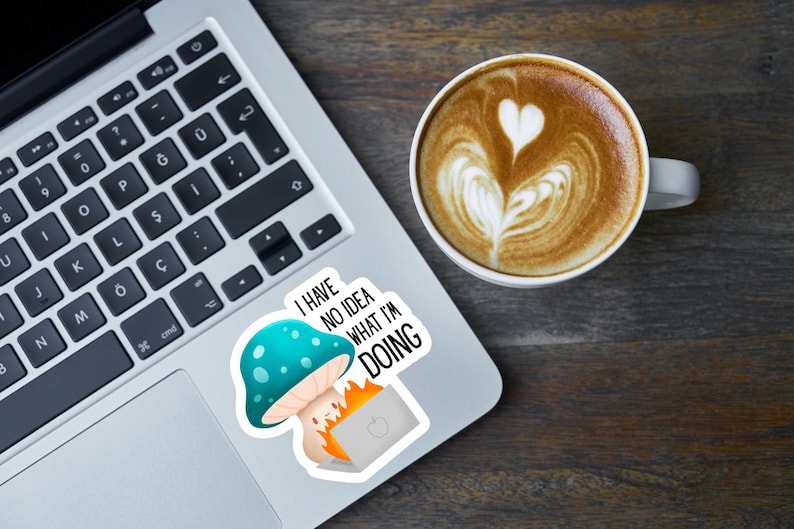 May include: A laptop with a sticker featuring a mushroom and a burning laptop, with the text "I have no idea what I'm doing." A cup of coffee with latte art is next to the laptop.