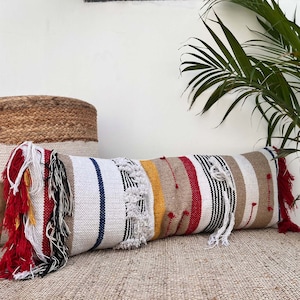 May include: A long, rectangular cushion with a white background and colourful stripes in red, yellow, black, and blue. The cushion has a fringe border and is made of woven fabric.