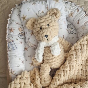 Könnte beinhalten: Eine beige gestrickte Decke mit einem beige gestrickten Teddybär, der einen weißen Schal trägt. Die Decke und der Teddybär befinden sich in einem weißen und blauen gemusterten Baby-Nest.