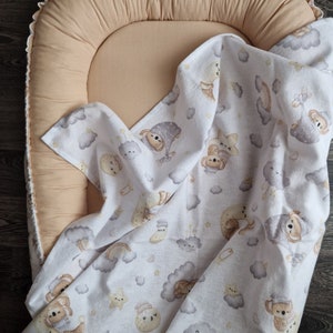 Op de afbeelding: Een wit babydekentje met een patroon van wolken, sterren en slapende beren. Het dekentje is gevouwen en ligt op een beige ovaalvormig baby nest.