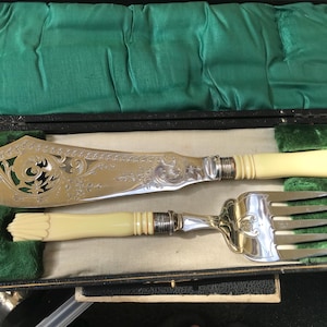 Puede incluir: Un juego de servir pescado plateado que consta de un cuchillo grande y ornamentado con calados y un tenedor a juego, ambos con mangos de color crema con detalles acanalados, presentados en un estuche negro forrado con tela verde y beige.