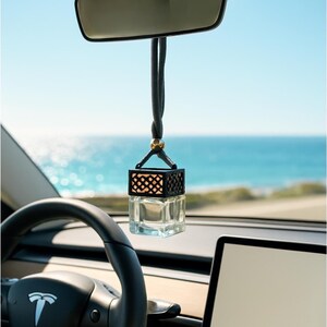Puede incluir: Un ambientador para coche colgado del espejo retrovisor. Tiene un frasco de vidrio transparente con un diseño de jaula geométrica negra. El fondo muestra un cielo azul brillante y el mar.