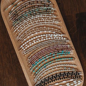 May include: A collection of beaded bracelets in various colors, including white, gold, turquoise, and black, displayed on a wooden tray. The bracelets feature small beads and gold accents, showcasing a variety of designs and styles.