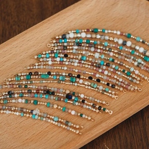 May include: A collection of beaded bracelets with gold-colored accents. The bracelets feature a variety of colorful beads, including turquoise, white, and pink, arranged in intricate patterns. The bracelets are displayed on a wooden tray.